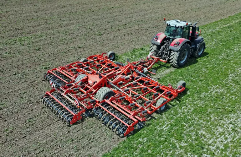 A red and gray tractor pulls Kuhn's tine cultivator Highlander with multiple disc rows across a brown agricultural field, with green crop rows visible alongside the working area.