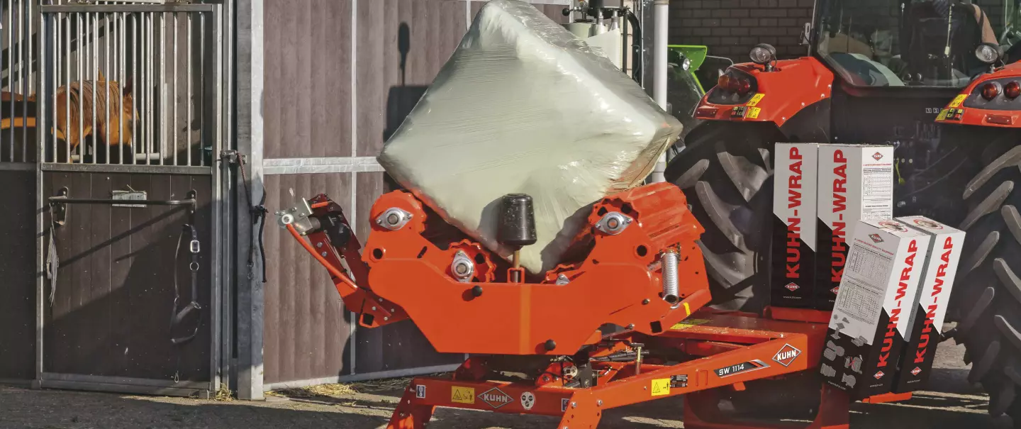 Une enrubanneuse pour balles rectangulaires SW1114 enrubannant une balle en position stationnaire devant une étable.