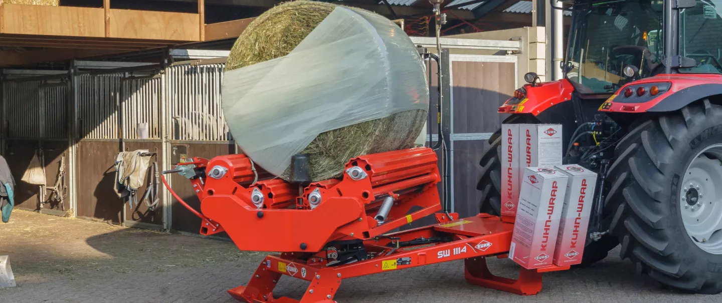 A KUHN SW1114 wrapping a round bale in front of a horse stable.